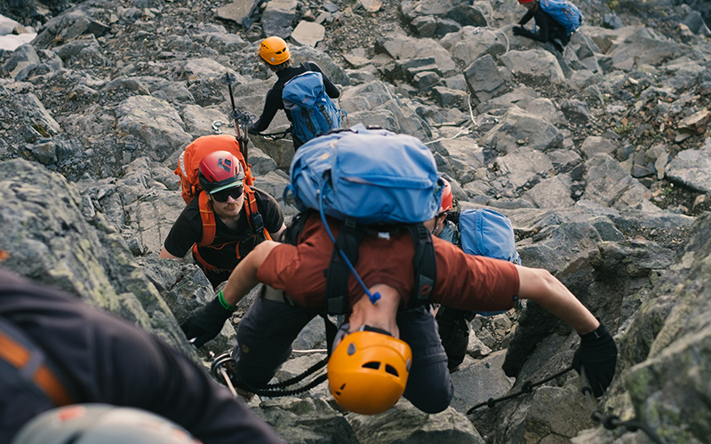 En grupp klättrare med hjälmar och stora ryggsäckar tar sig uppför en brant, stenig fjällsida med hjälp av rep.