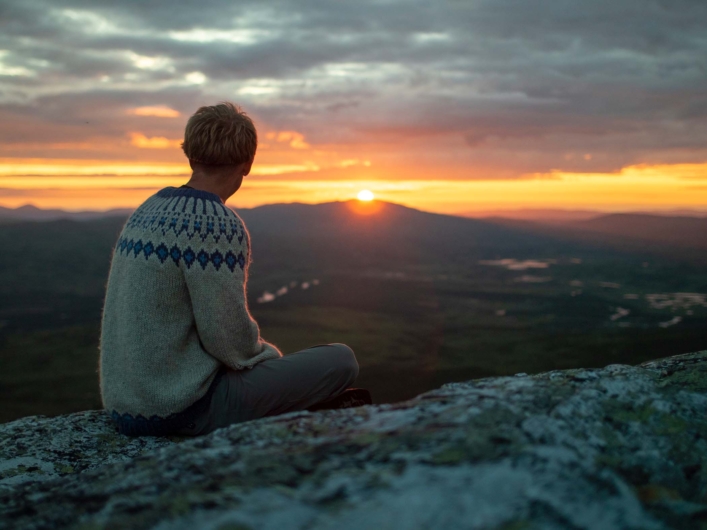 Person sitter på en klippa och blickar ut över midnattssol. 