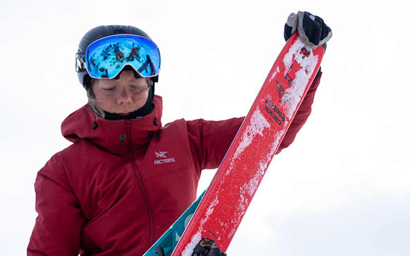 En skidåkare i röd jacka och hjälm inspekterar en snötäckt skida i vintermiljö.