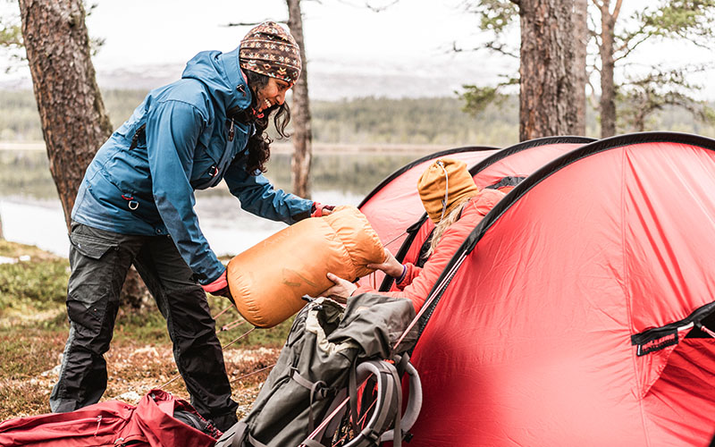 En person räcker över en packad sovsäck till en annan som sitter vid öppningen av ett rött tält i skogsmiljö vid en sjö.