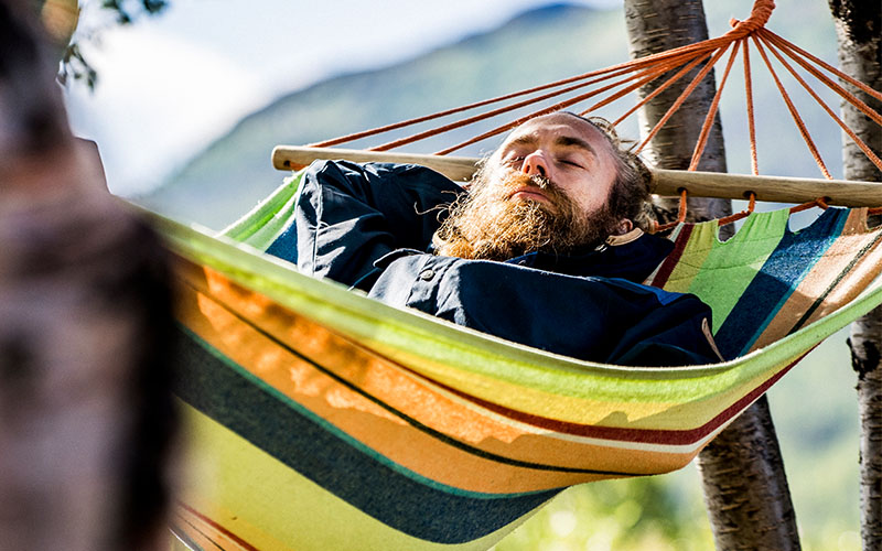 En person ligger och vilar i en färgglad hängmatta mellan två träd i soligt fjälllandskap.