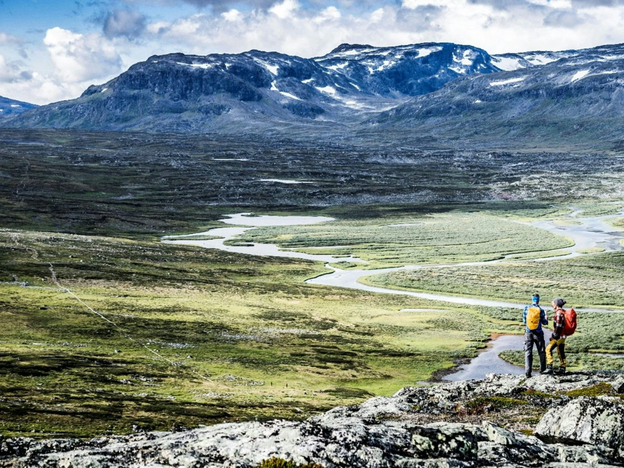 Två vandrare med ryggsäckar blickar ut över ett vidsträckt fjällandskap med slingrande vattendrag och höga berg i bakgrunden.