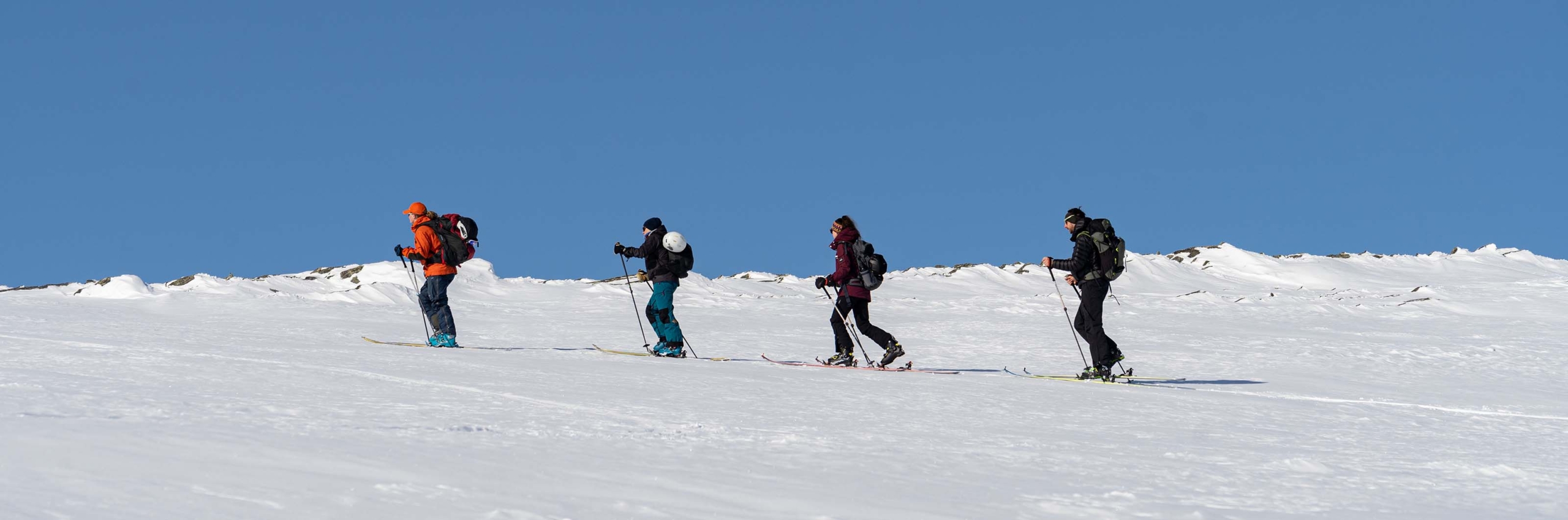 Fyra skidåkare går på turskidor uppför en snötäckt fjällrygg under klarblå himmel.
