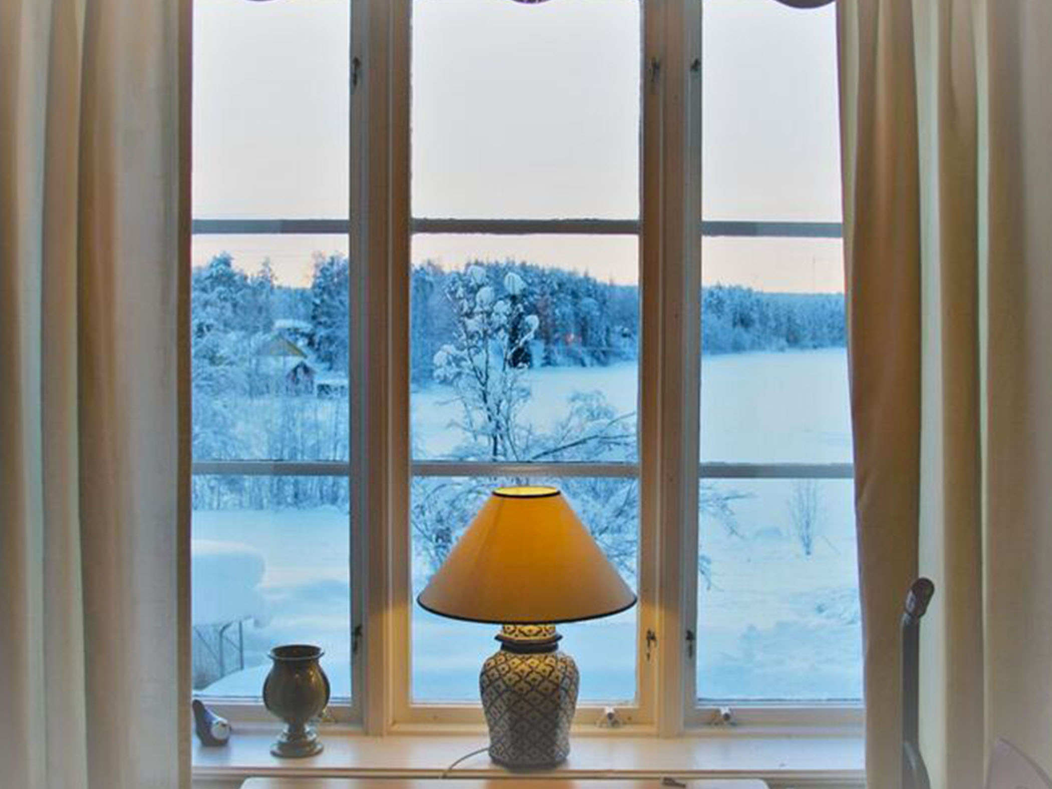 Fönster med tänd bordslampa och utsikt över ett snötäckt vinterlandskap vid Jädraås herrgård.