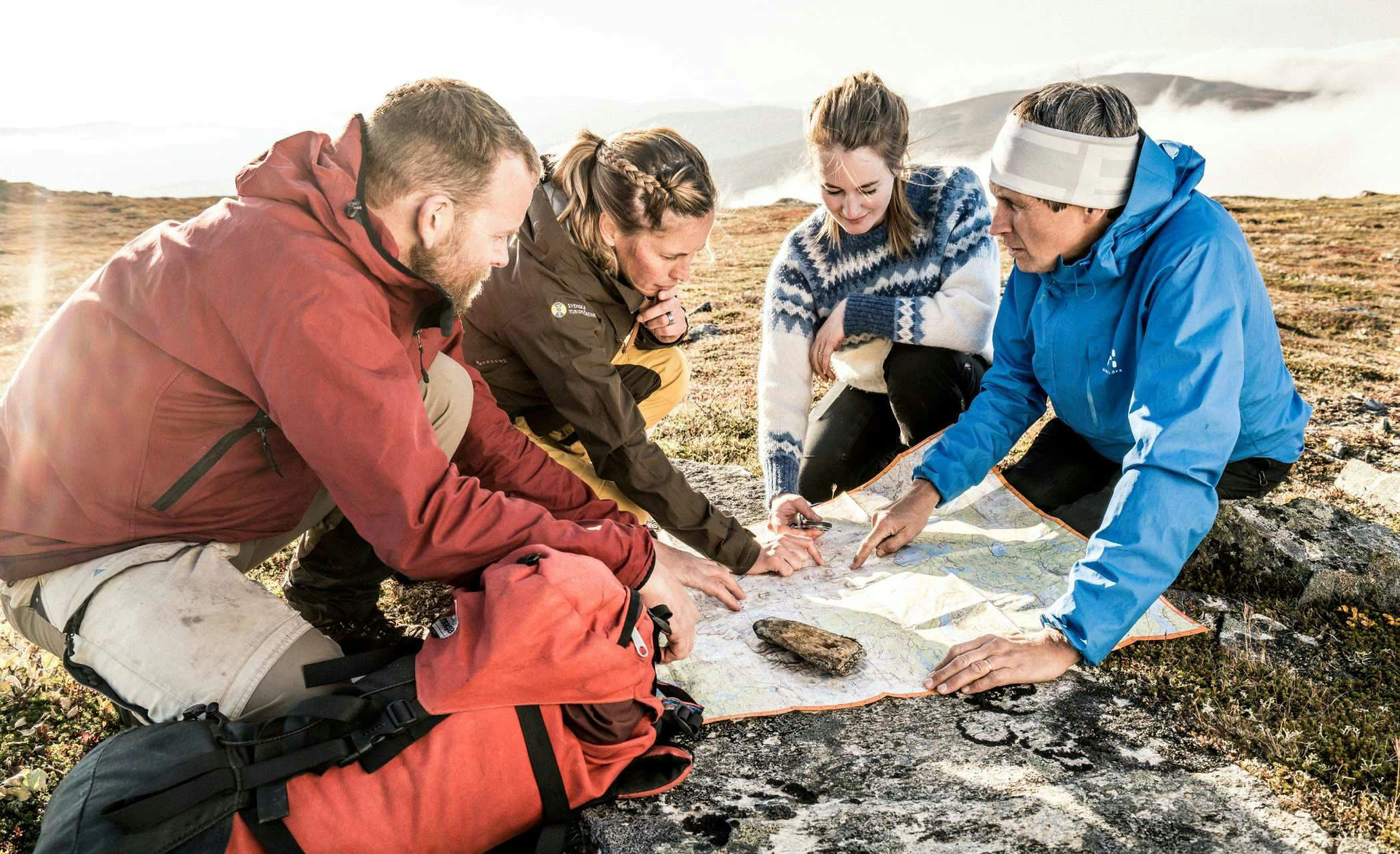En grupp människor studerar en karta tillsammans på en stenig fjällsluttning i soligt väder.
