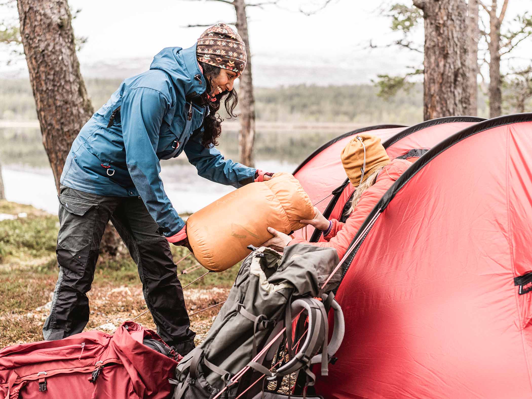 En person räcker över en packad sovsäck till en annan som sitter vid öppningen av ett rött tält i skogsmiljö vid en sjö.