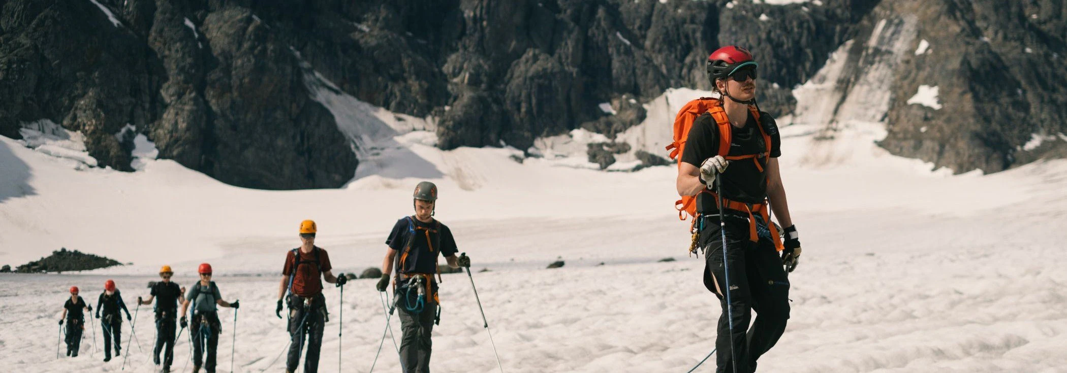 En grupp klättrare med hjälmar och sele vandrar på en glaciär, ledda av en guide. Höga klippor och snö syns i bakgrunden.