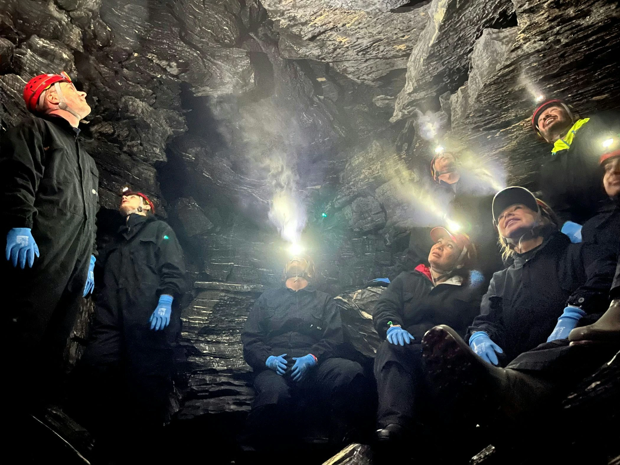 En grupp personer iförda overaller, hjälmar med pannlampor och handskar sitter och står inne i en mörk grotta med glänsande stenväggar. Ljuset från pannlamporna lyser upp den trånga grottan.
