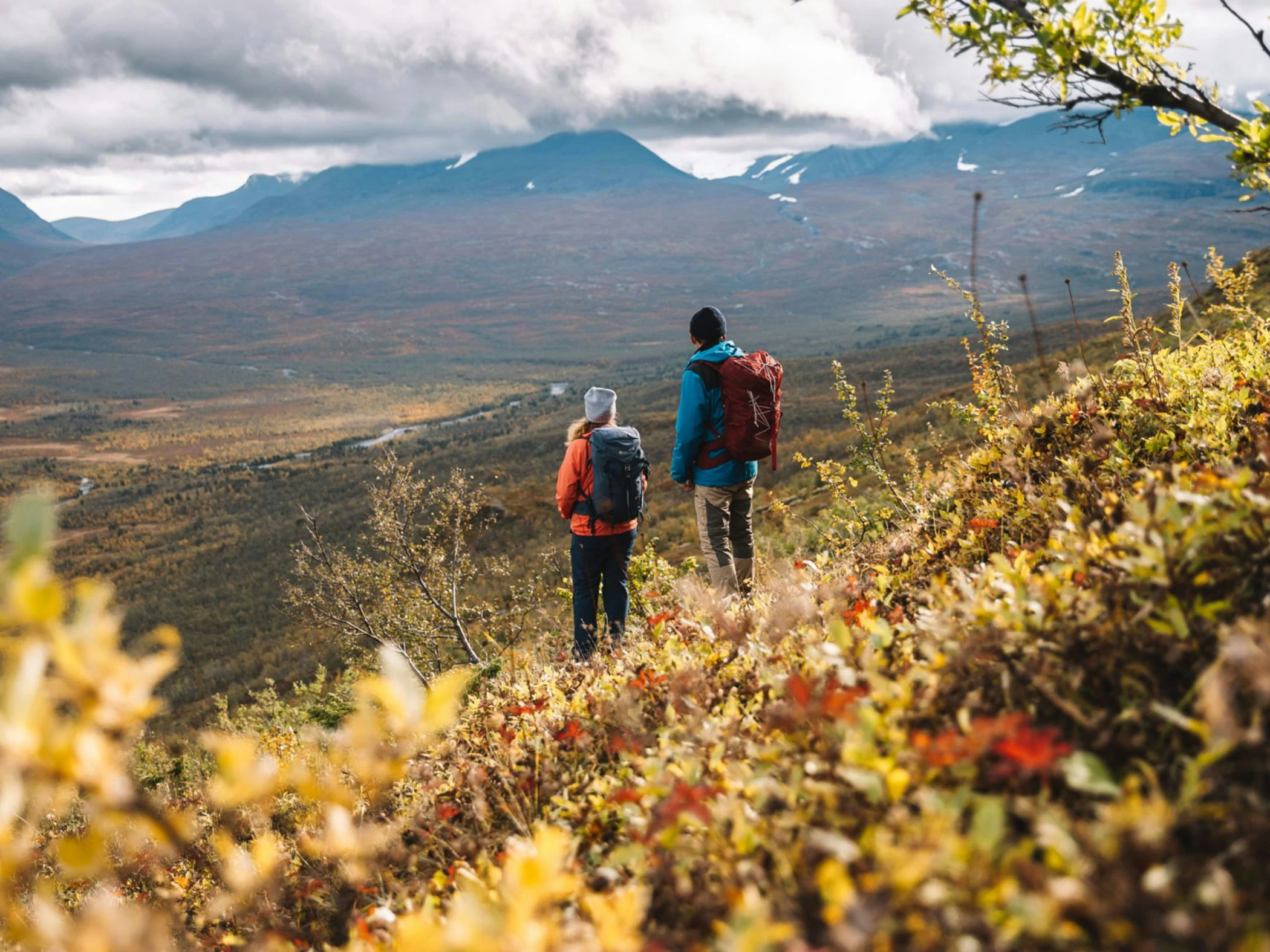 Två vandrare med ryggsäckar står på en sluttning täckt av höstfärger och blickar ut över en dal med fjäll i bakgrunden. Himlen är molnig och landskapet breder ut sig under dem.
