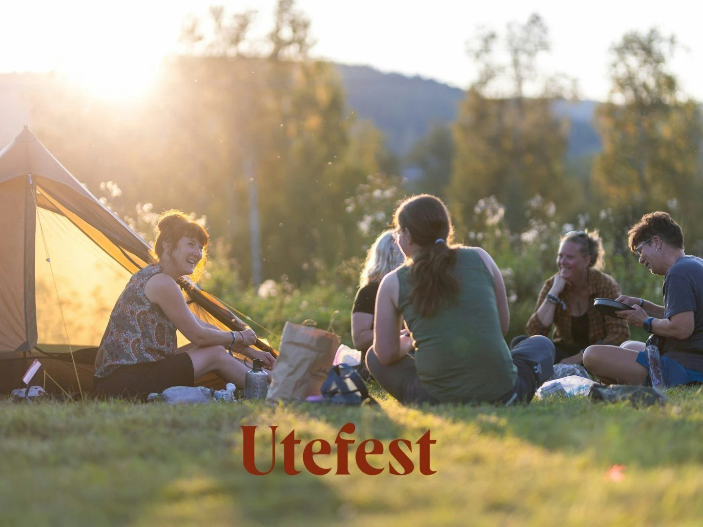 En grupp människor sitter i en cirkel på en gräsmatta vid solnedgången, med ett tält i bakgrunden.
