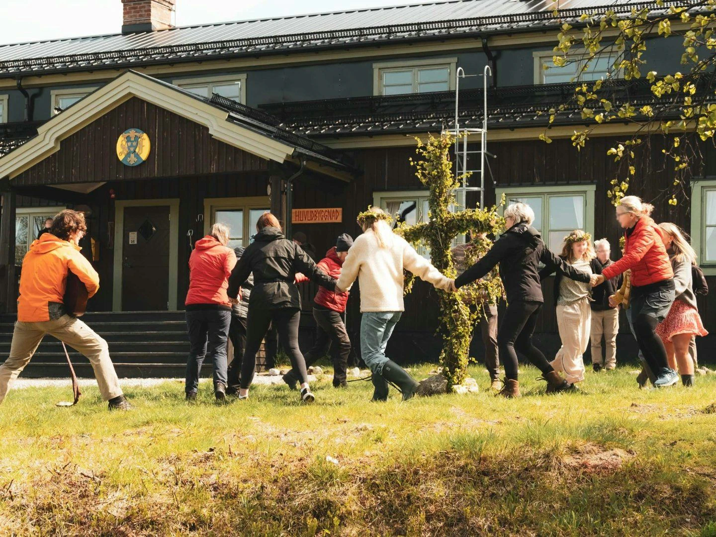 Människor dansar runt en midsommarstång framför en träbyggnad i solsken.