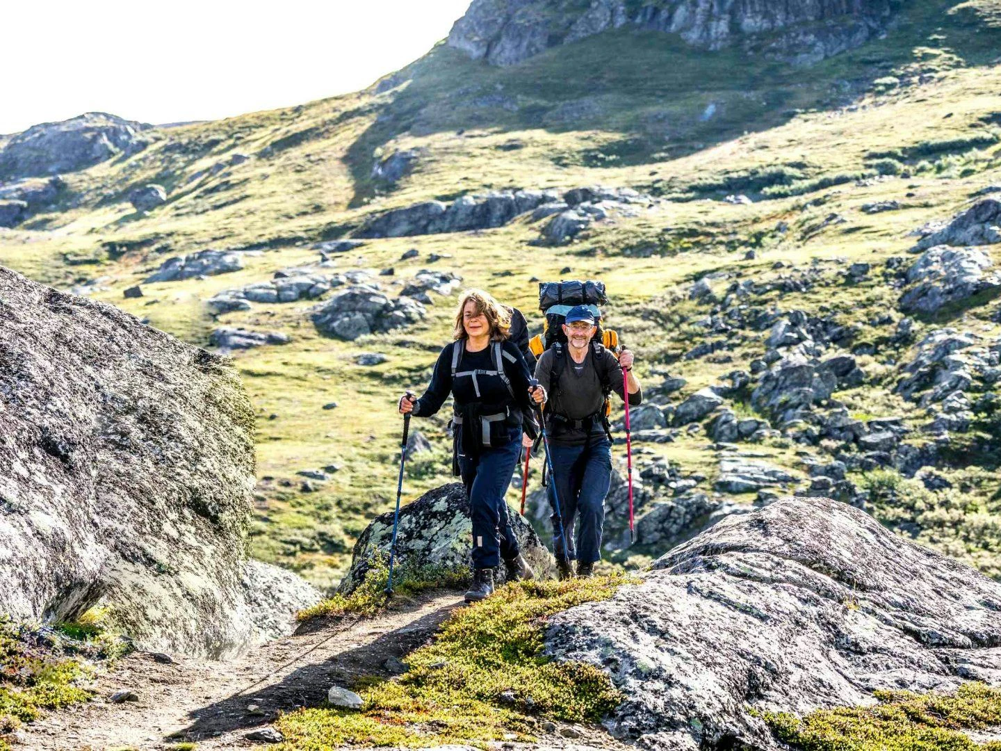 Två vandrare med ryggsäckar och vandringsstavar går på en stig i en bergig och grönskande miljö.