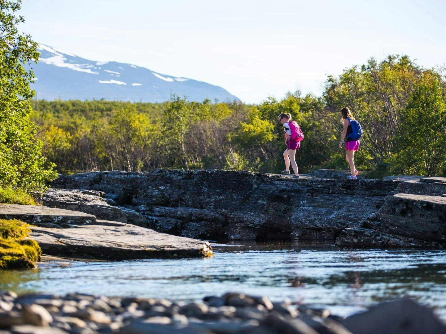 Två personer vandrar över klippor nära en bäck med berg och skog i bakgrunden.
