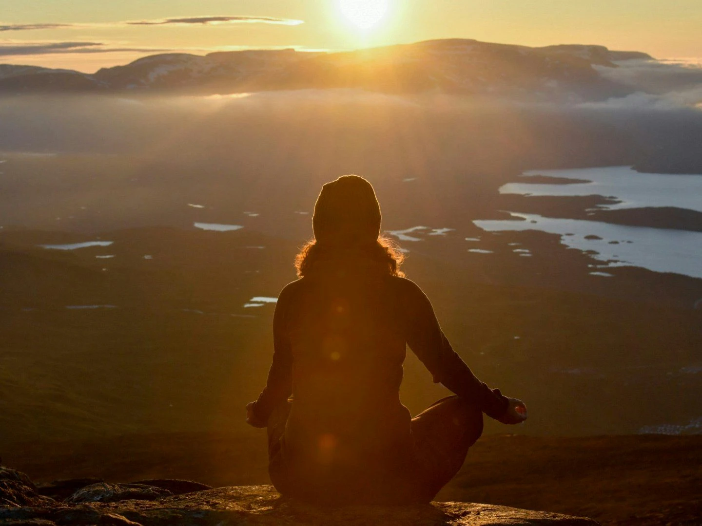 En person sitter i meditationsställning på en bergstopp vid solnedgång, med utsikt över sjöar och berg i fjärran.