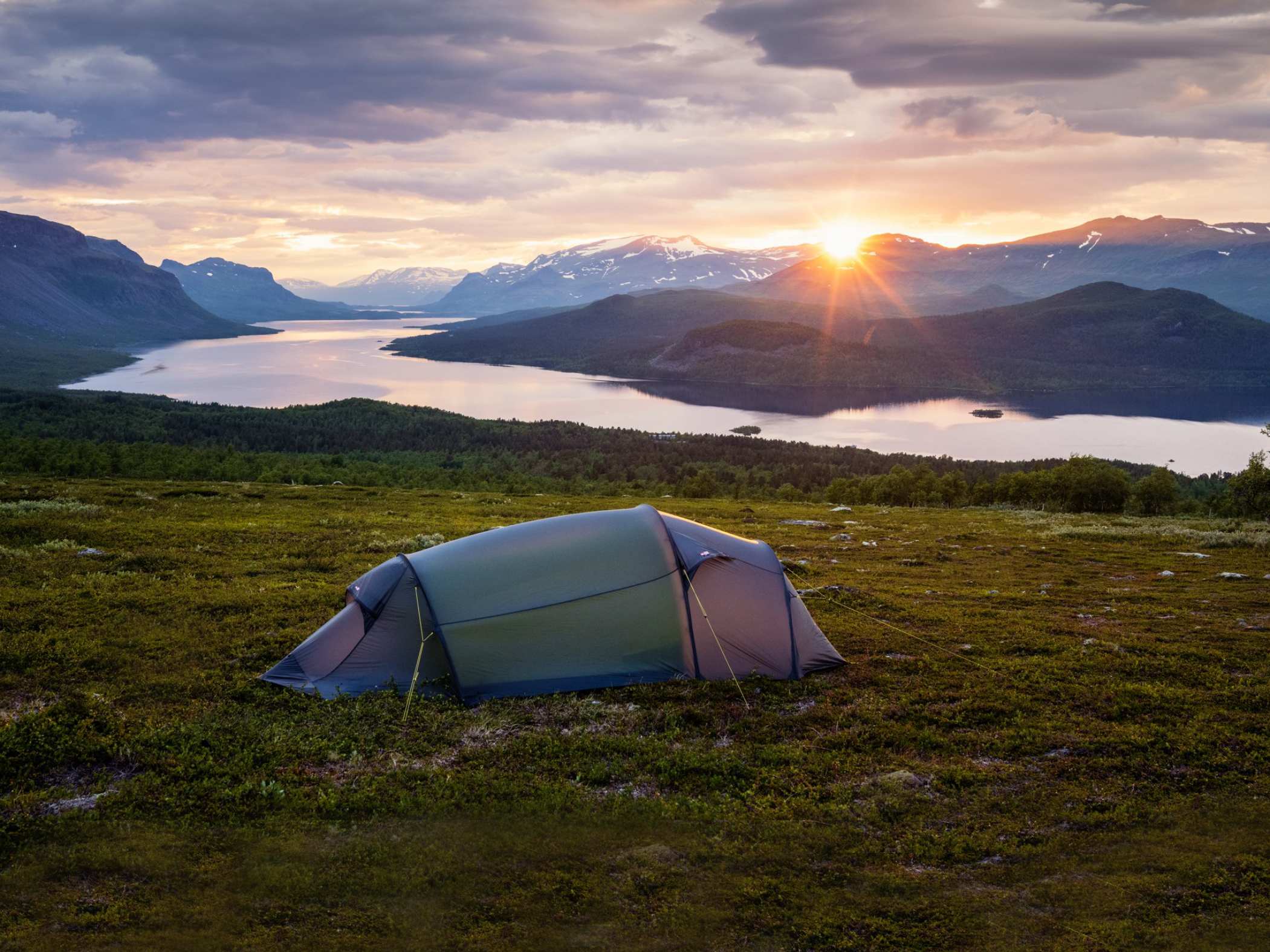 Ett tält står på en grön fjälläng med utsikt över en sjö omgiven av berg. Solen håller på att gå ner bakom bergen, och himlen är färgad i mjuka toner av orange, rosa och lila.