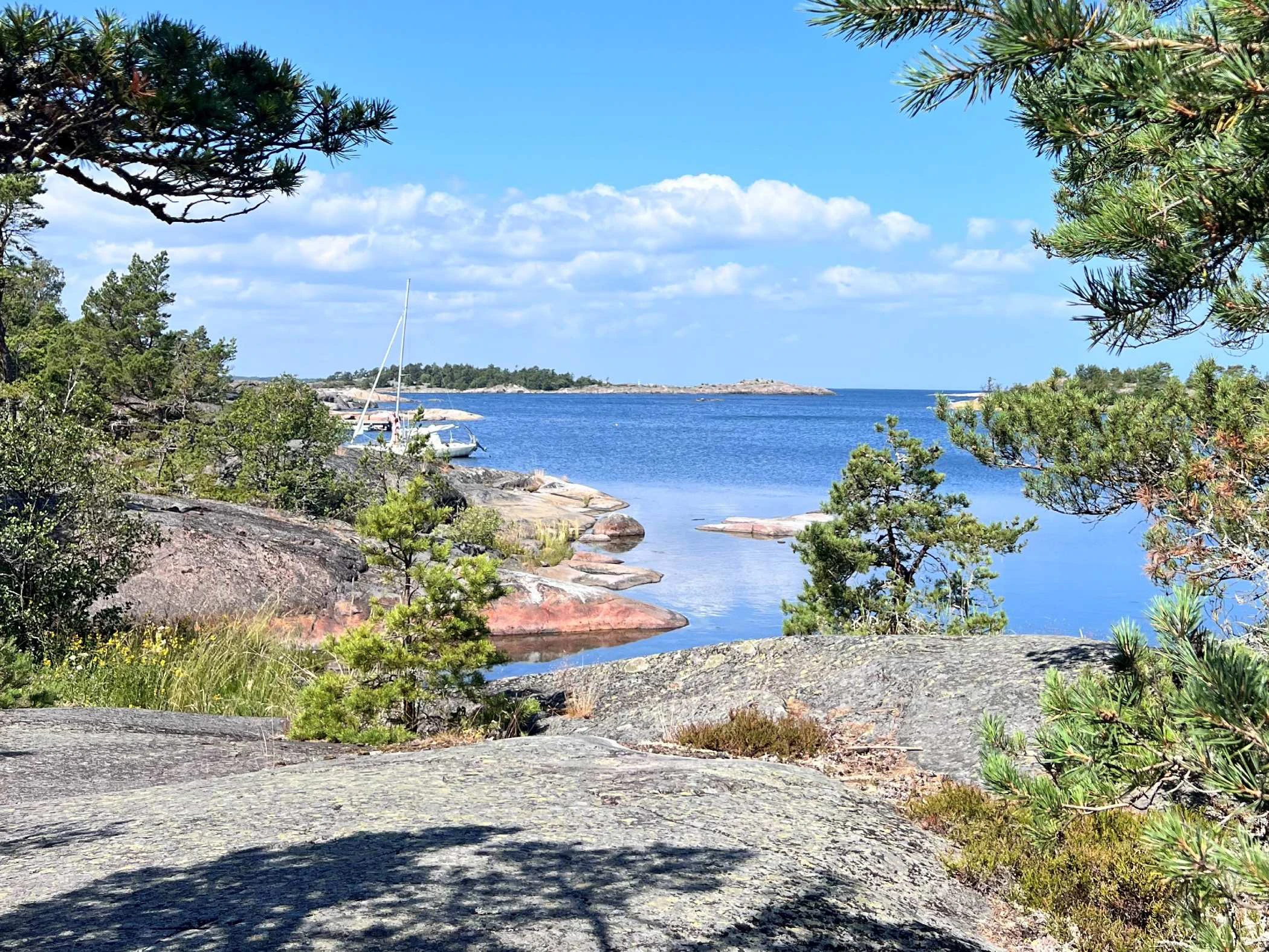 En solig skärgårdsvy med klippor, tallar och lugnt blått hav, där en segelbåt ligger förtöjd vid land.