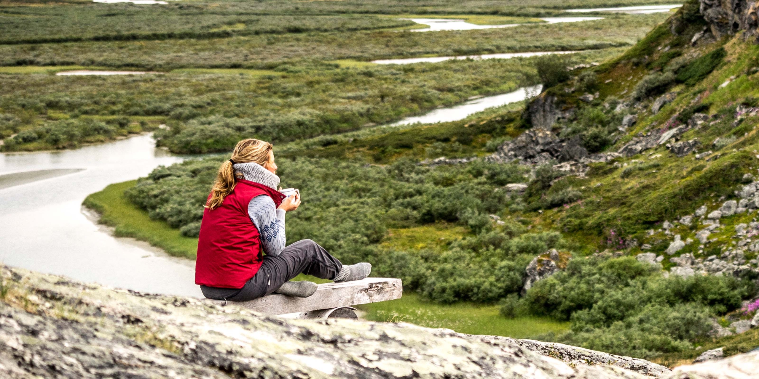 En person sitter på en bänk och dricker ur en kopp med utsikt över ett grönt fjällandskap med slingrande vattendrag.