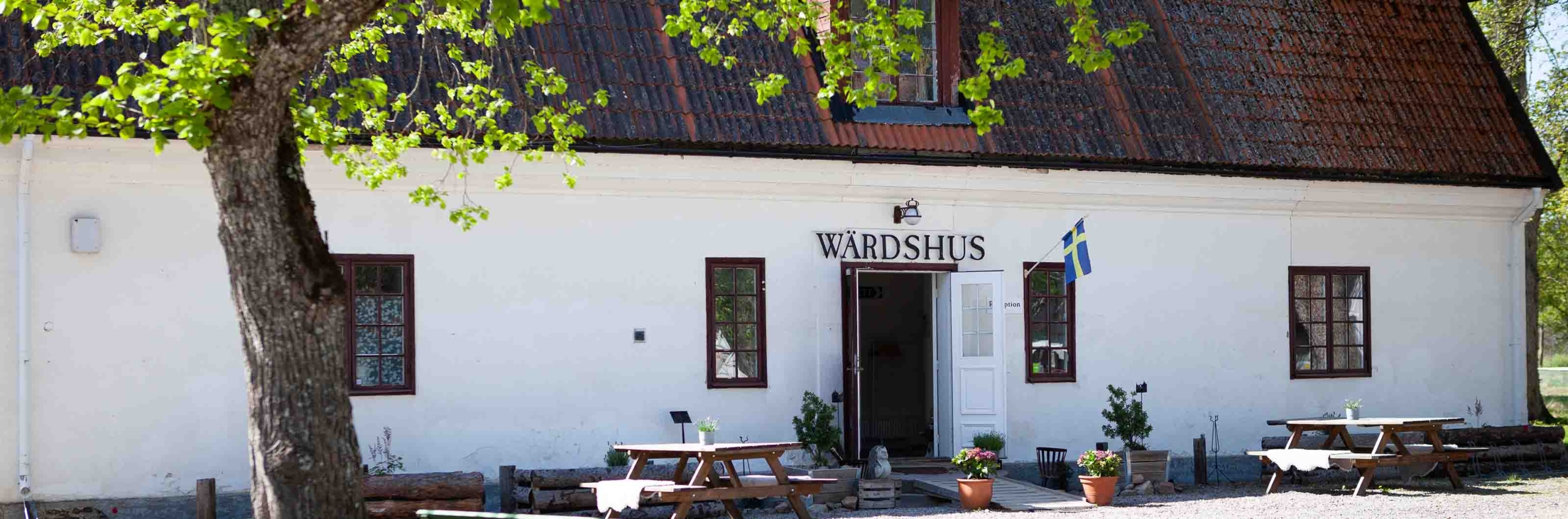 Värdshus med vita väggar och uteservering i grönskande miljö.