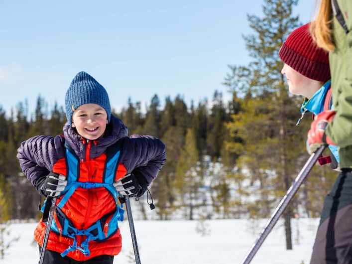 Leende barn på skidor i snöigt landskap, iklädd färgglada vinterkläder och ryggsäck, med skog i bakgrunden och ett annat barn bredvid.