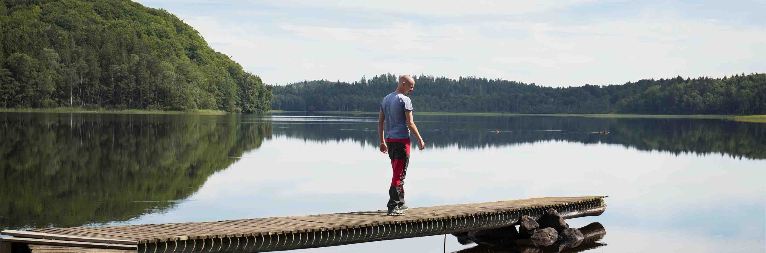 Person står på en brygga vid en spegelblank sjö omgiven av skog.