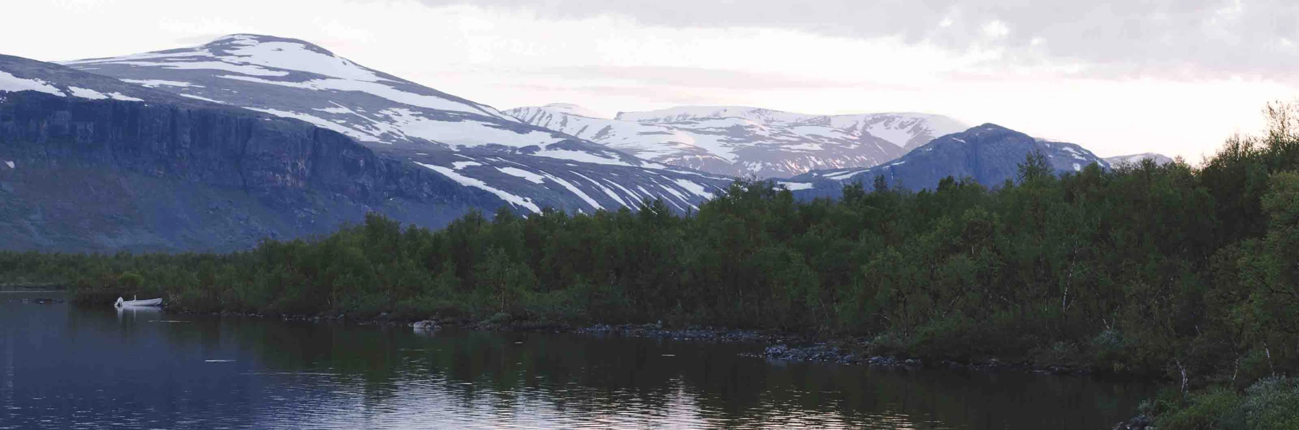 Fjällandskap med sjö och snöklädda berg under molnig himmel.