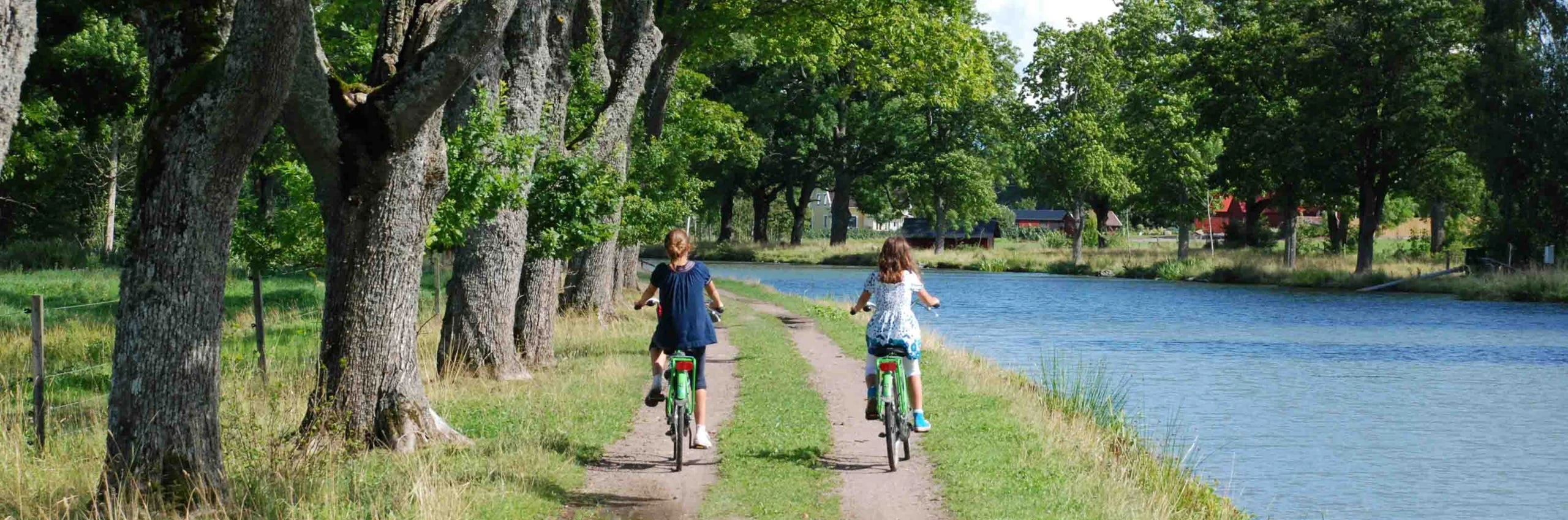 Två barn cyklar längs en grusväg kantad av träd, med en kanal som glittrar bredvid.