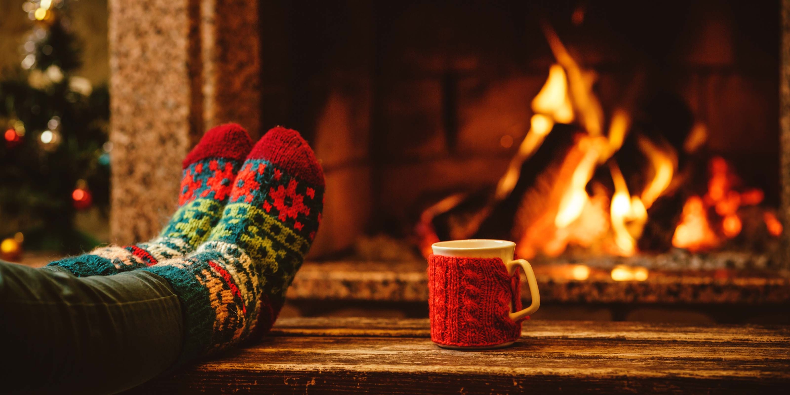 Fötter i stickade sockor vilar framför en sprakande brasa med en röd mugg på golvet intill.