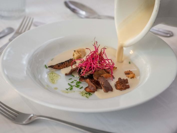 Krämig soppa hälls upp i en djup tallrik med svamp, krutonger, örter och färska groddar som garnering.