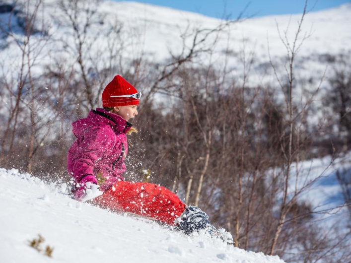 Ett barn i färgglada vinterkläder åker nerför en snötäckt backe i fjällmiljö, med kala björkar och snöklädda sluttningar i bakgrunden.