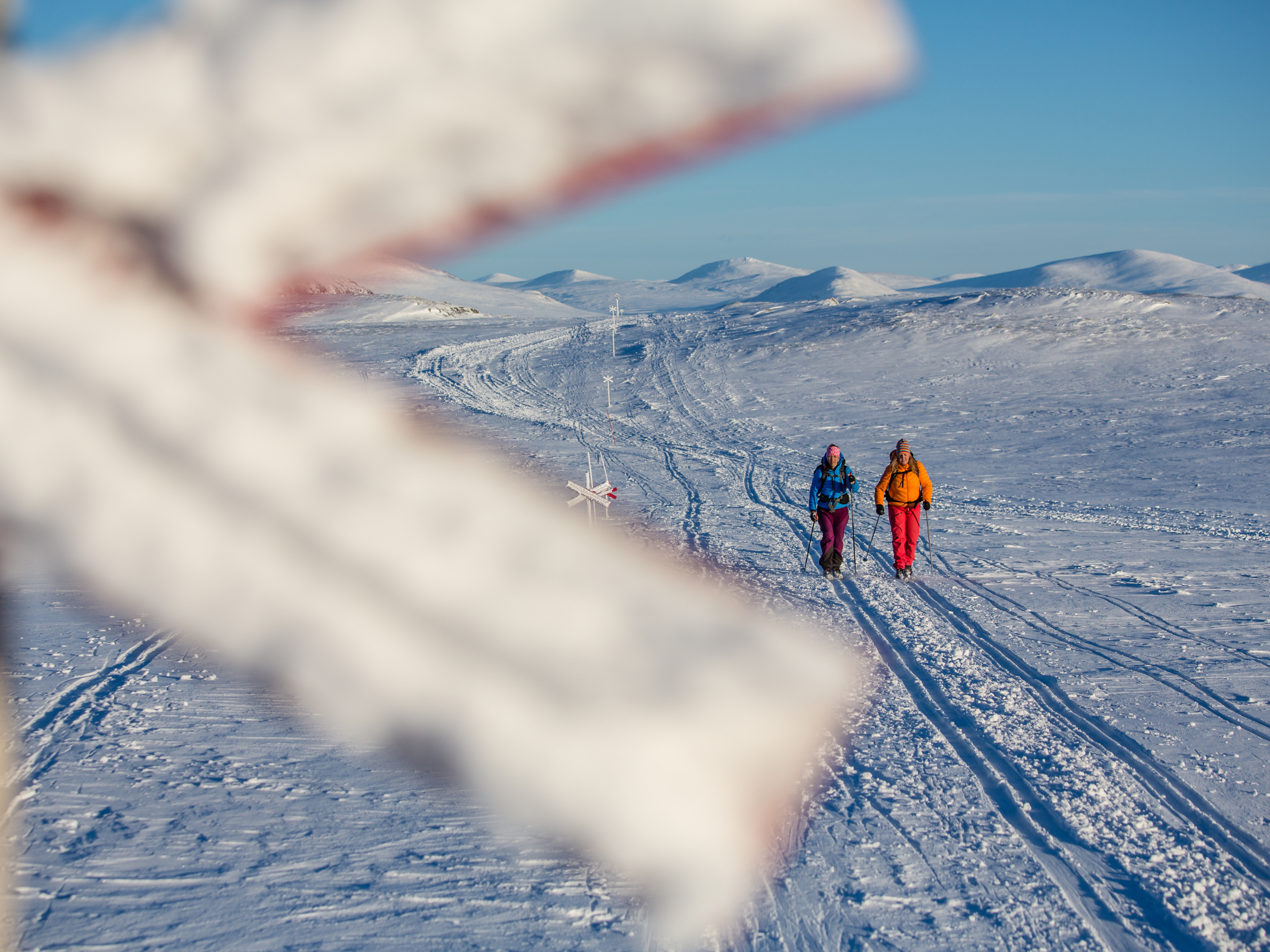Två personer vandrar med stavar över ett snötäckt fjälllandskap i vinterljus.