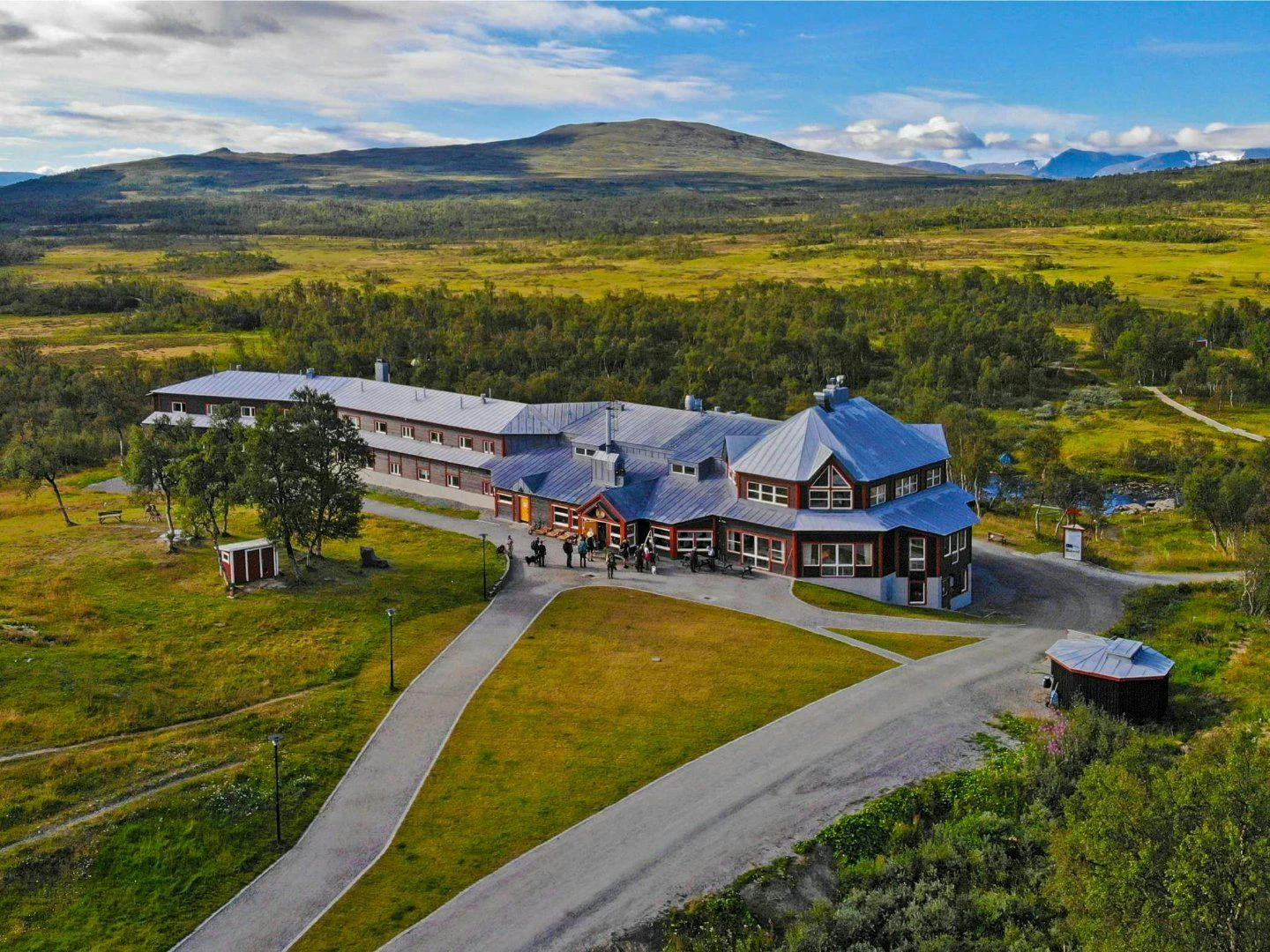 Flygbild av STF Storulvån Fjällstation, en stor byggnad med röd träfasad och vita detaljer, omgiven av grönskande fjällandskap. I bakgrunden syns vidsträckta fjäll och en blå himmel med moln. En asfalterad väg leder fram till fjällstationen.