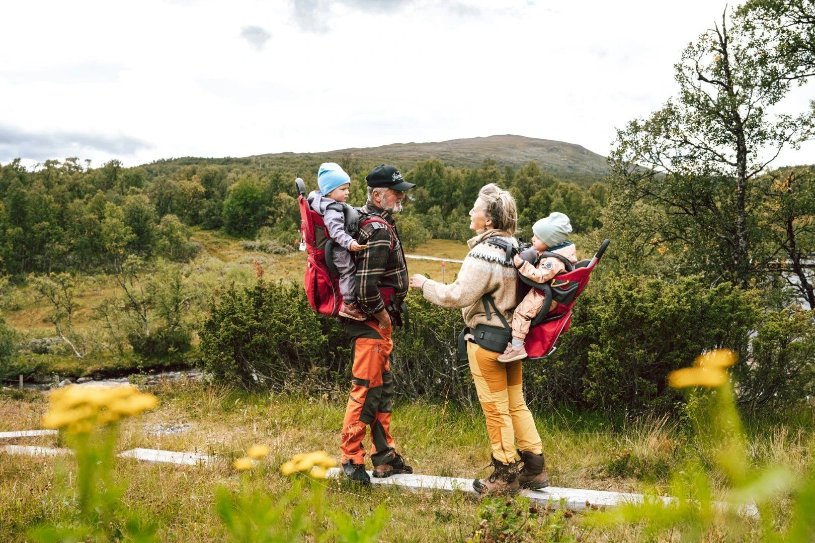 Två vuxna bär varsitt barn i bärstol på ryggen. De står på en stig omgiven av grönska, buskar och berg i bakgrunden. Bilden har en varm och lugn känsla med inslag av gula blommor i förgrunden.