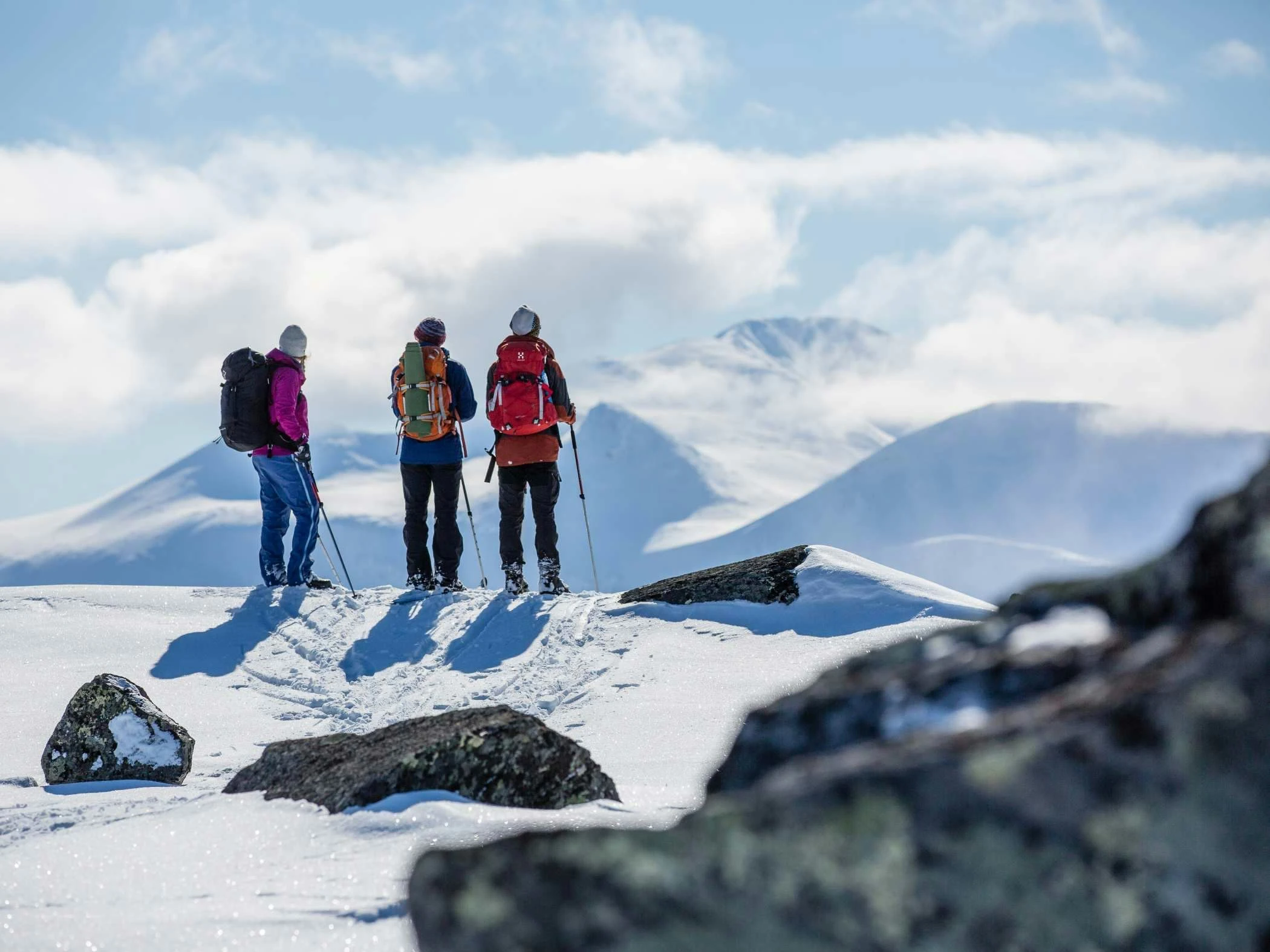 Tre personer står med ryggsäckar och vandringsstavar på ett snötäckt fjäll, med en bergskedja och moln i bakgrunden under en klarblå himmel. Stenar syns i förgrunden.