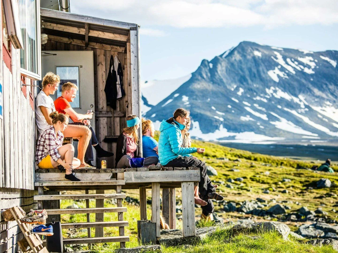 En grupp människor sitter på en träveranda utanför en stuga i fjällmiljö, omgivna av grönska och berg med snöfläckar i bakgrunden. Solen skiner och de verkar njuta av utsikten och umgänget i en avkopplad atmosfär.