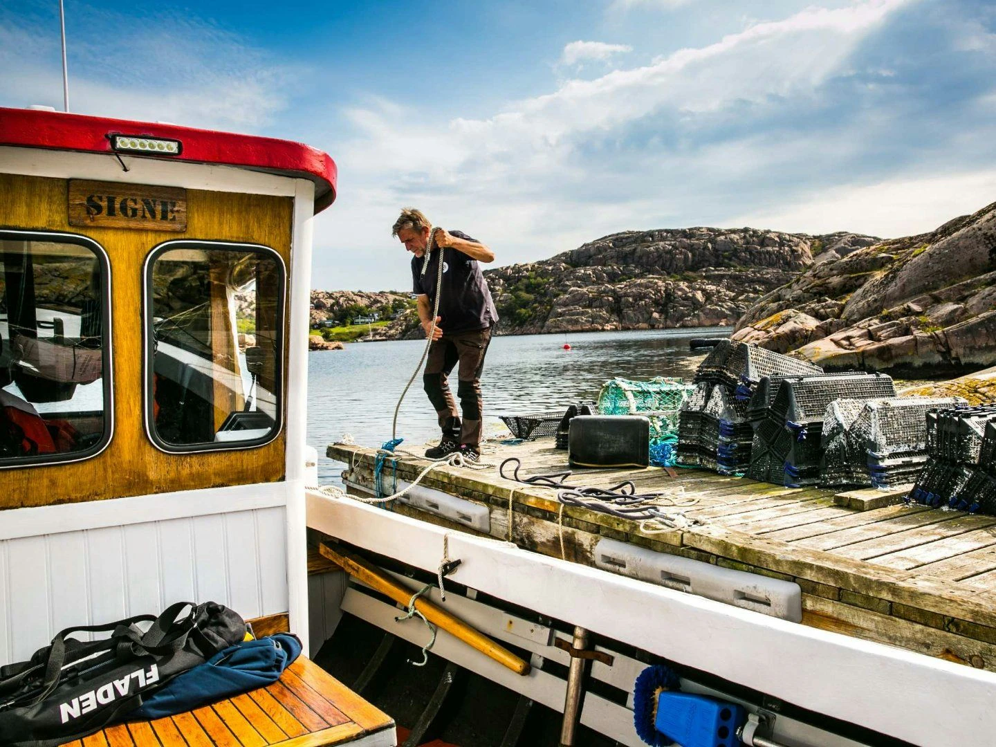 Man på brygga drar i ett rep bredvid en båt, med hummertinor staplade på bryggan och klippor i bakgrunden.