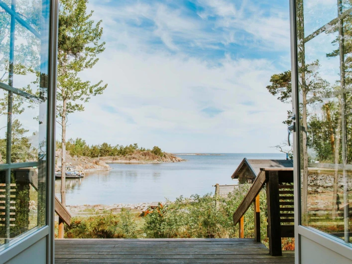 Utsikt över lugn skärgård från öppen dörr, med träd, klippor och en liten stuga vid vattnet.