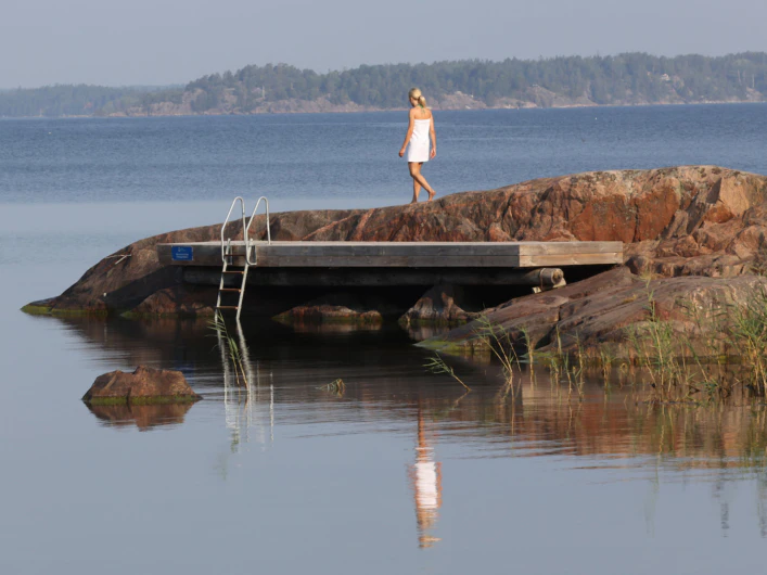 Kvinna i vit handduk går på klippor vid lugnt vatten, intill en liten badbrygga med stege.