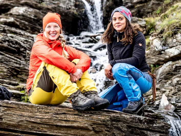Två vandrare sitter på en klippa framför ett litet vattenfall i fjällmiljö, klädda i friluftskläder med ryggsäck bredvid.