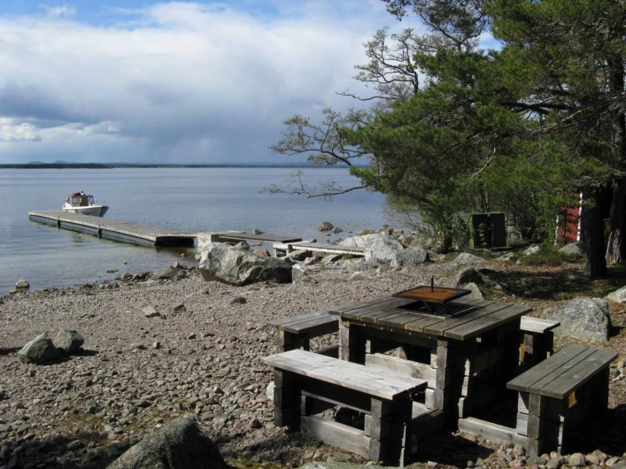 Stenig strand med brygga, båt och picknickbord vid lugnt hav.