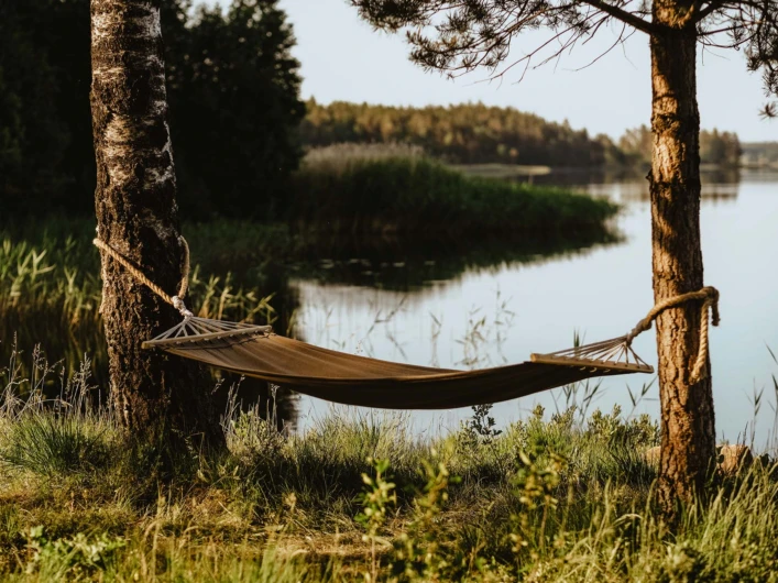Hängmatta uppspänd mellan två träd vid en stilla sjö, omgiven av grönska.
