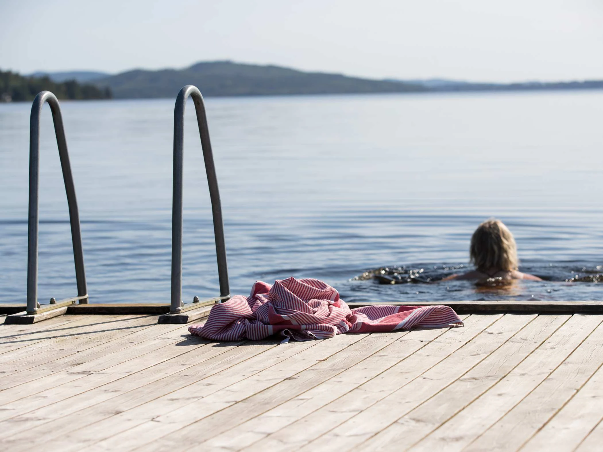 En träbrygga vid en sjö, med en badstege till vänster. En person simmar i vattnet i bakgrunden, och ett rosa tygstycke ligger på bryggan i förgrunden.