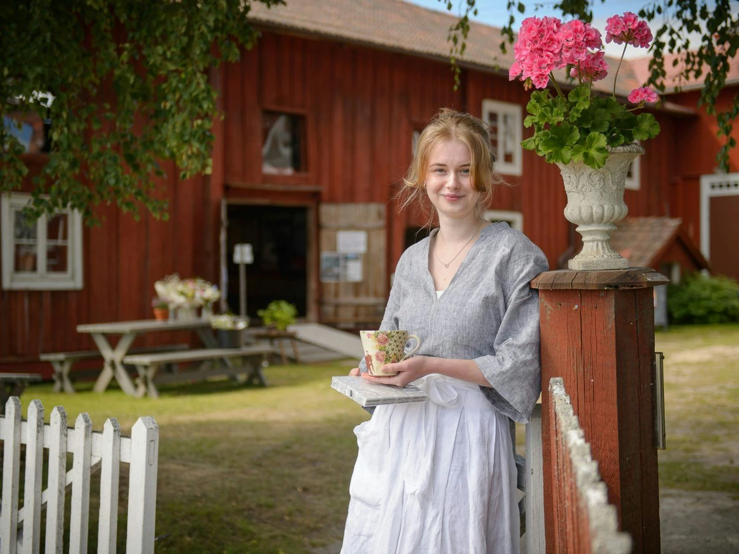 Person står vid ett staket med en kopp och bok, framför en röd träbyggnad med blommor.