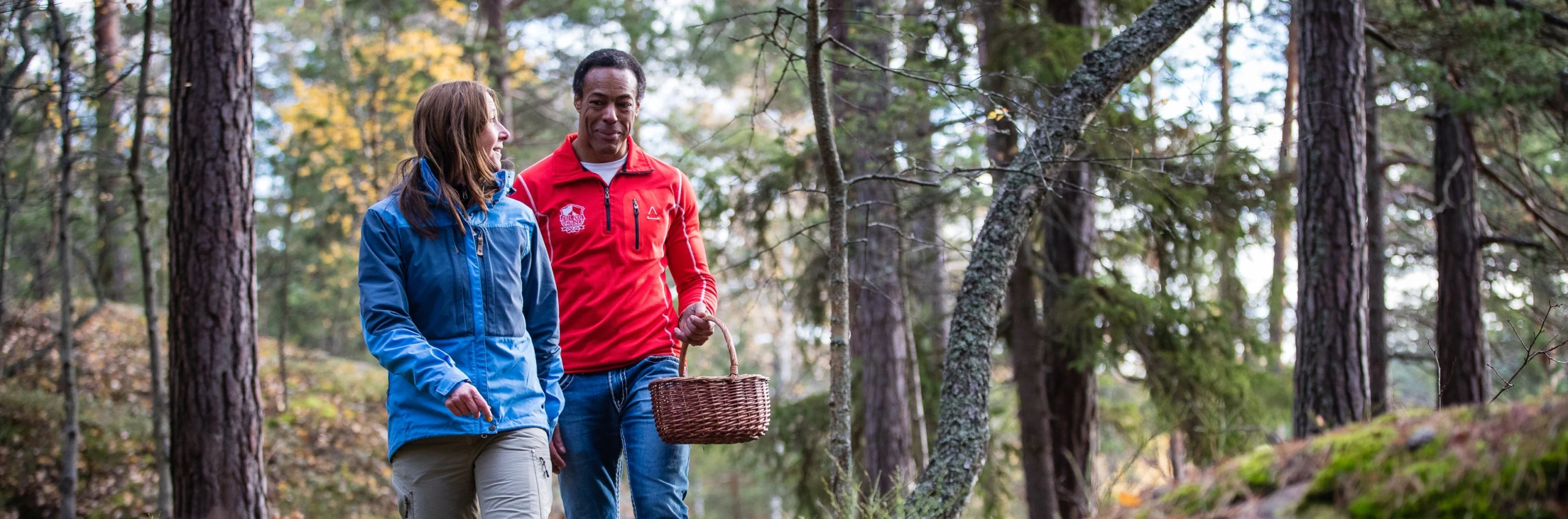 Två personer vandrar i skogen med svampkorg.