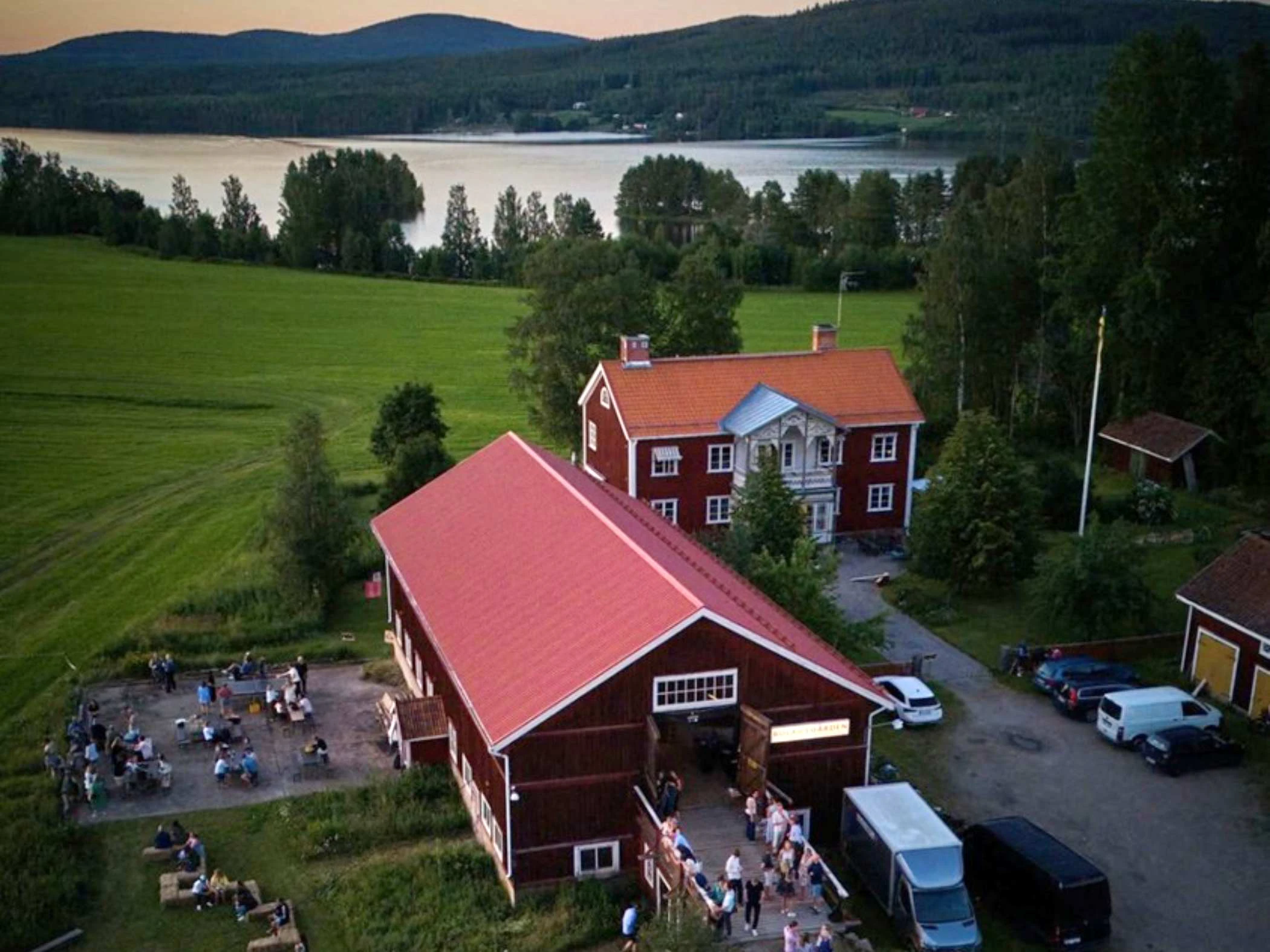 En flygbild över två röda hus och en äng och en sjö samt berg i bakgrunden.