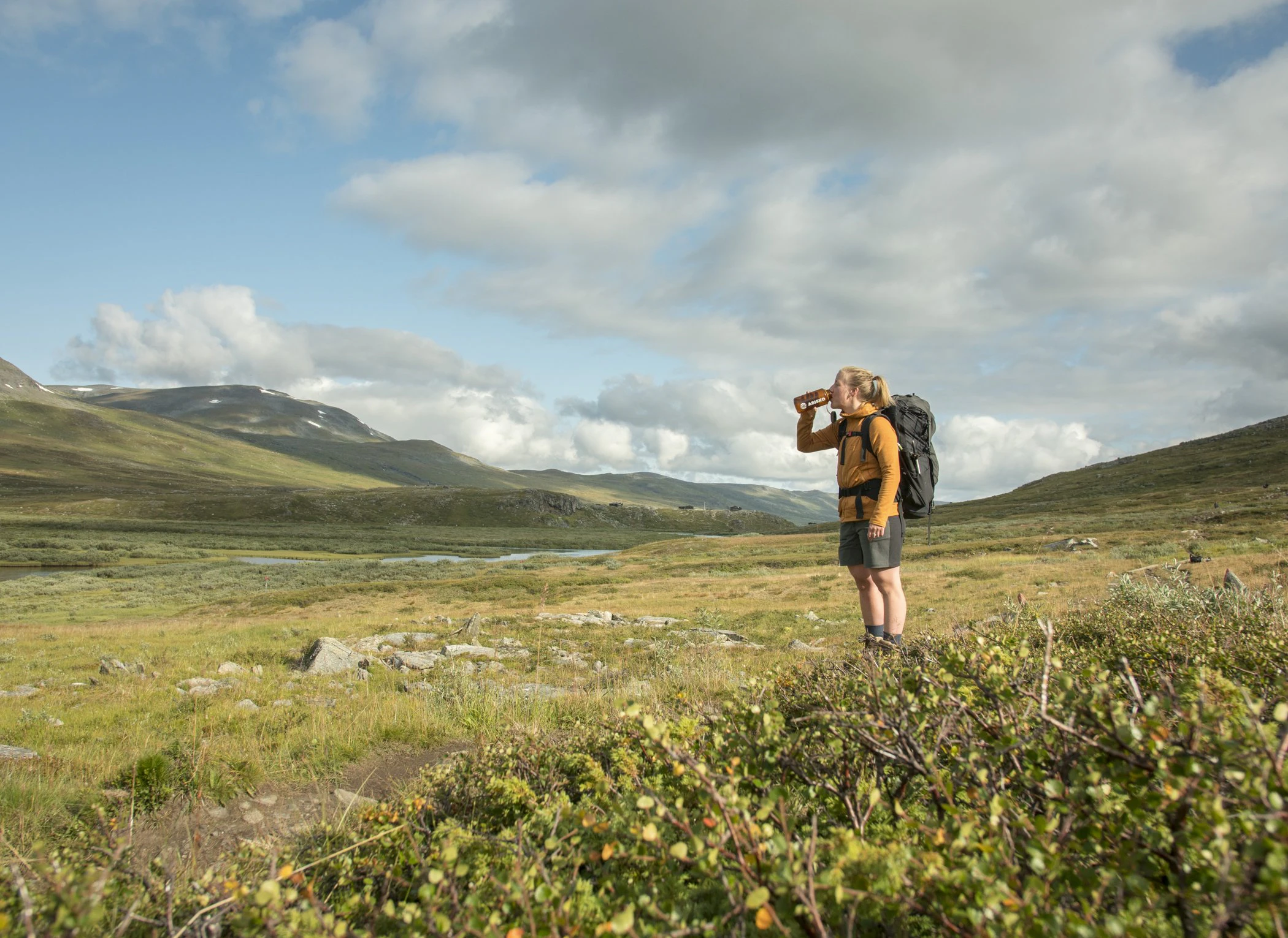 Vandrare med ryggsäck dricker ur flaska i öppet fjällandskap.
