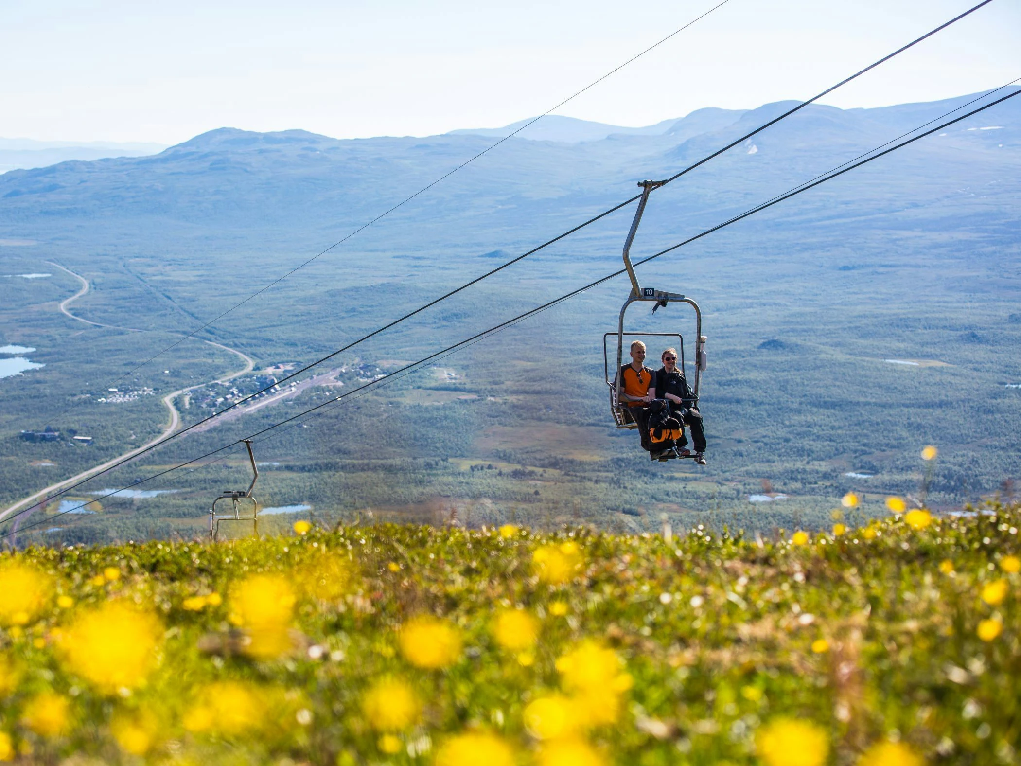 Två personer åker sittlift över berget Nuolja med gula blommor i förgrunden.