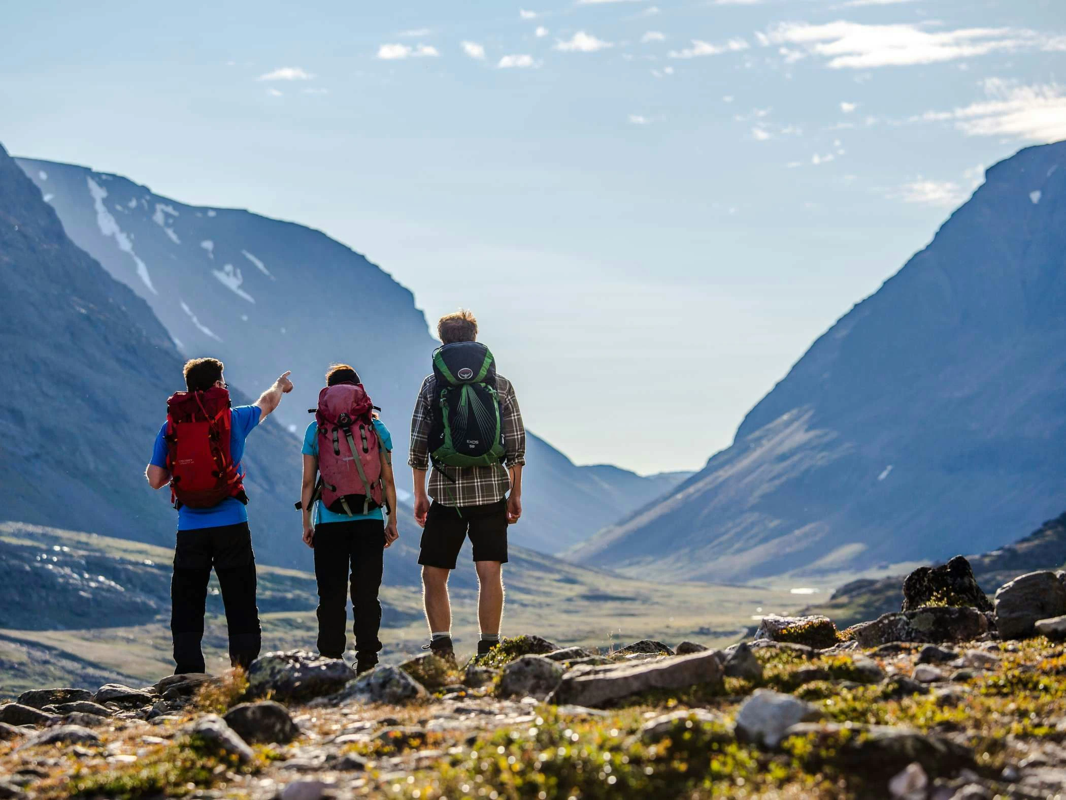 Tre vandrare med ryggsäckar står på en stenig fjällsluttning och blickar ut över en dal omgiven av höga berg. En av dem pekar mot horisonten under en klarblå himmel.