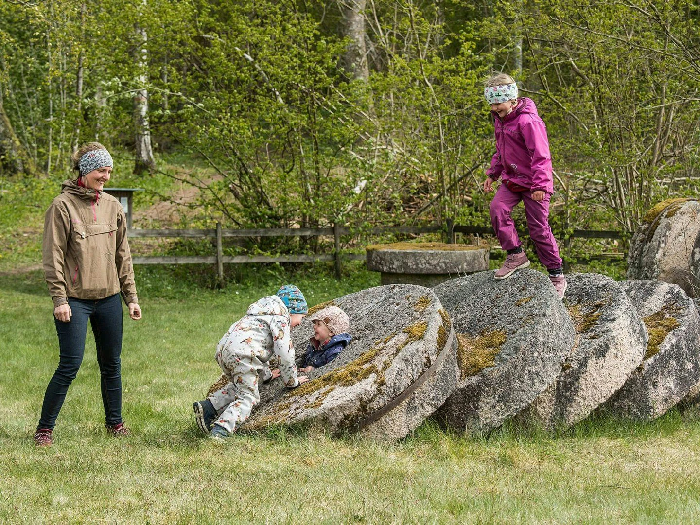 Barn leker klättrande på stora mossbelupna kvarnstenar medan en vuxen ser på leende.