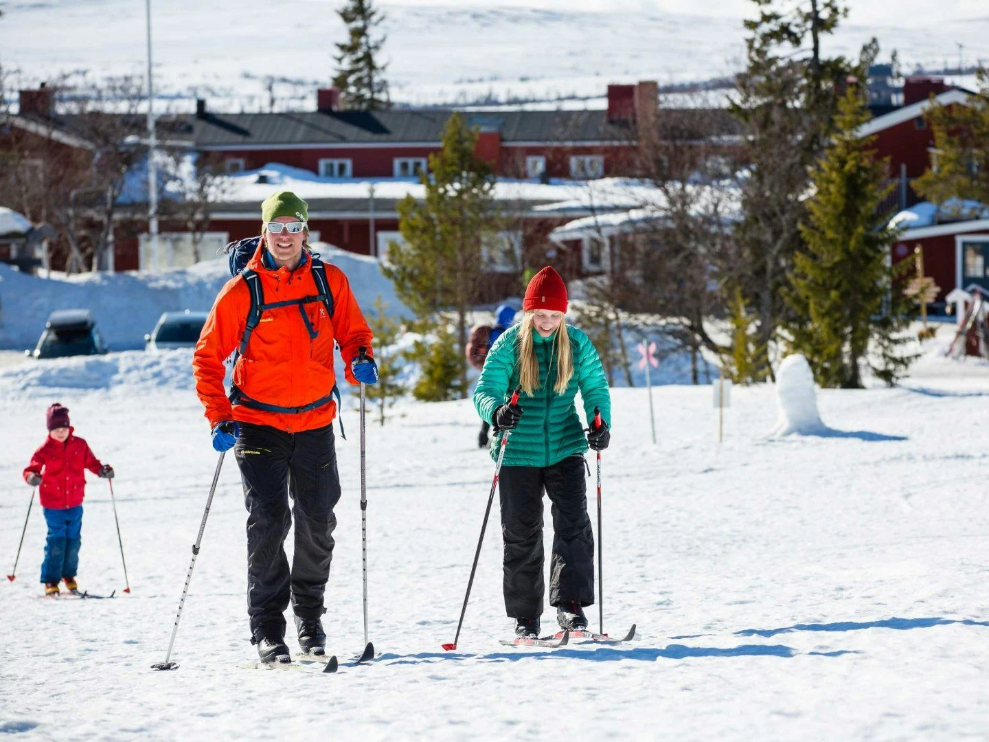 En pappa i röd jacka och två barn som åker skidor framför ett rött hus.
