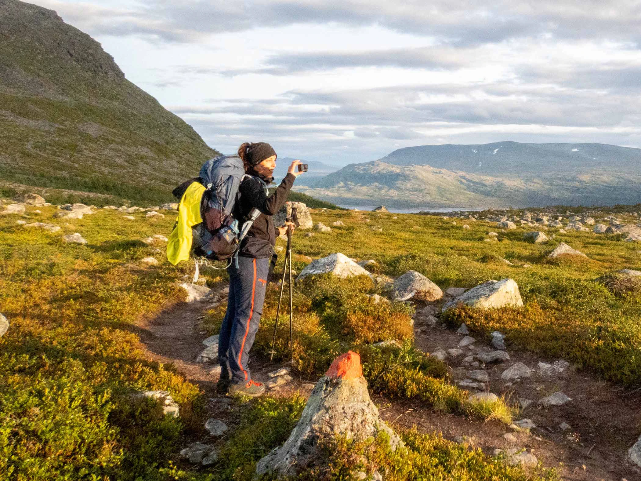 En person med stor ryggsäck står på en fjällstig och fotograferar det vidsträcka landskapet. Runt omkring syns låga fjällväxter, stenar och berg i kvällsljus under en molnig himmel.